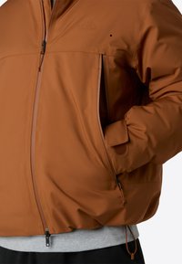 Braune Outdoor-Jacke mit glatter Textur, Reißverschluss vorne und seitlichen Taschen. Verfügt über eine verstellbare Kordel auf Taille und ein dezentes Logo.