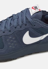 Tmavě modrá teniska z broušené kůže a síťoviny, se bílým znakem Nike, texturovanými tkaničkami a odolnou gumovou podrážkou s jemnými perforacemi.