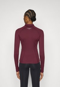 Bordo krāsas garām piedurknēm aktīvs tops ar augstu apkakli, ar pieguļošu dizainu, plakaniem šuvēm un logotipu augšējā muguras daļā. Mīksts, elastīgs audums.