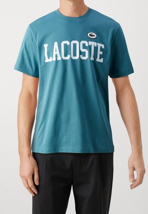 Turkusowy T-shirt z krótkim rękawem z wyrazistym białym napisem "LACOSTE" oraz okrągłą metką logo na klatce piersiowej. Wykonany z miękkiego materiału bawełnianego.