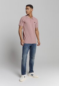 Lichtroze polo shirt met korte mouwen, kraag, drie knopen en een klein logo. Draag het met een blauwe spijkerbroek en witte sneakers.