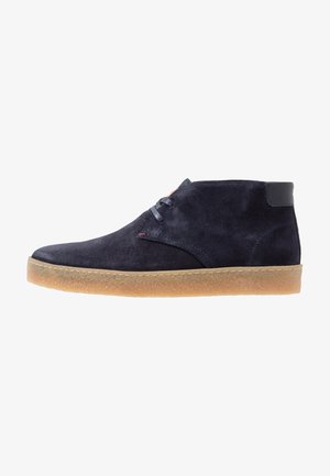 Chaussure cheville en daim bleu marine avec deux œillets pour lacets, semelle en caoutchouc naturel et pièce de cuir noir au talon, vue de côté.