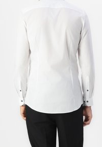 Camicia bianca a maniche lunghe con design aderente, texture liscia e dettagli in bottone neri sui polsini, indossata sopra pantaloni neri.