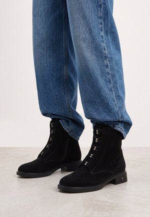 Person trägt blaue Jeans und schwarze geschnürte Stiefeletten und steht auf einem hellgrauen Boden.