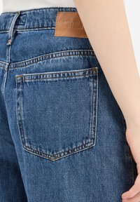 Donkere spijkerbroek van denim, met een leren label "DRYKORN," een achterzak en contrasterende stiksels langs de naden.