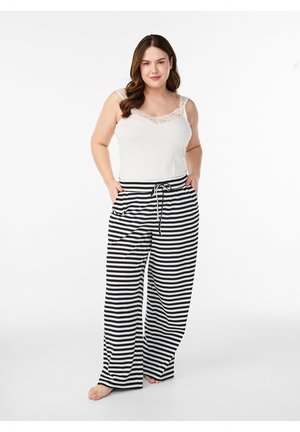 Woman standing barefoot wearing a white lace-trimmed tank top and black-and-white horizontal striped wide-leg pants.