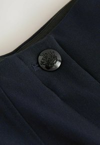 Musta metallipainike kohokuviolla varustetulla pääkallosymboolilla tummansinisessä teksturoidussa kankaassa, korostaen vaatteen yksityiskohtia ja muotoilua.