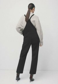 Schwarze Latzhose mit verstellbaren Trägern, getragen über einem hellgrauen Pullover. Das Outfit verfügt über weite Hosenbeine und braune Stiefel mit Absatz.