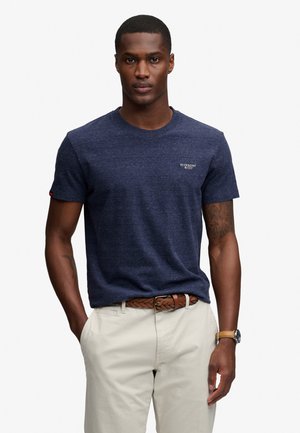 Man draagt marineblauw T-shirt met korte mouwen, beige broek, gevlochten bruine riem en horloge, staat met één hand in de zak tegen een witte achtergrond.
