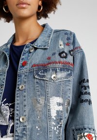 Džínová bunda se stříbrnými flitrovými písmeny, vyšívanými hvězdami, květinami a červeným prošíváním, nošena přes tmavě modrou košili s bílým grafickým potiskem.