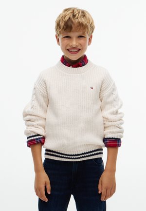 Ragazzo biondo sorridente che indossa un maglione bianco a maglia con orlo a righe e maniche di camicia rossa a quadri arrotolate, in piedi davanti a uno sfondo semplice.