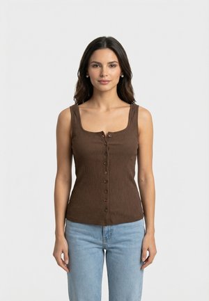 Donna con capelli scuri che indossa un top marrone senza maniche con bottoni e jeans azzurri chiari, in piedi davanti a uno sfondo chiaro e uniforme.