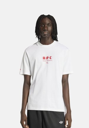 Personne avec des dreadlocks mi-longs portant un T-shirt blanc AFC Holloway Rd London et un short noir Adidas sur un fond uni.