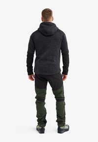 Man wearing dark gray hooded jacket and green and black outdoor pants, standing facing away on a white background.