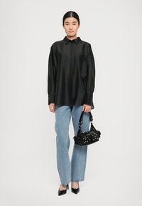Svart sateng skjorte med lange ermer og knapper på kragen, kombinert med lys blå rette jeans. Modellen holder en liten svart håndveske med nagler.