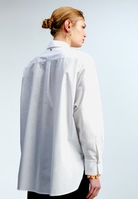 Camicia bianca oversized con maniche lunghe, polsini con bottone e piega sul retro. Presenta un piccolo logo "M" dorato al colletto. Texture liscia.
