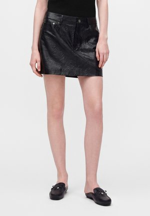 Personne portant une mini-jupe en cuir texturé noire, un haut noir sans manches et des mocassins noirs sans lacets, debout devant un fond uni.