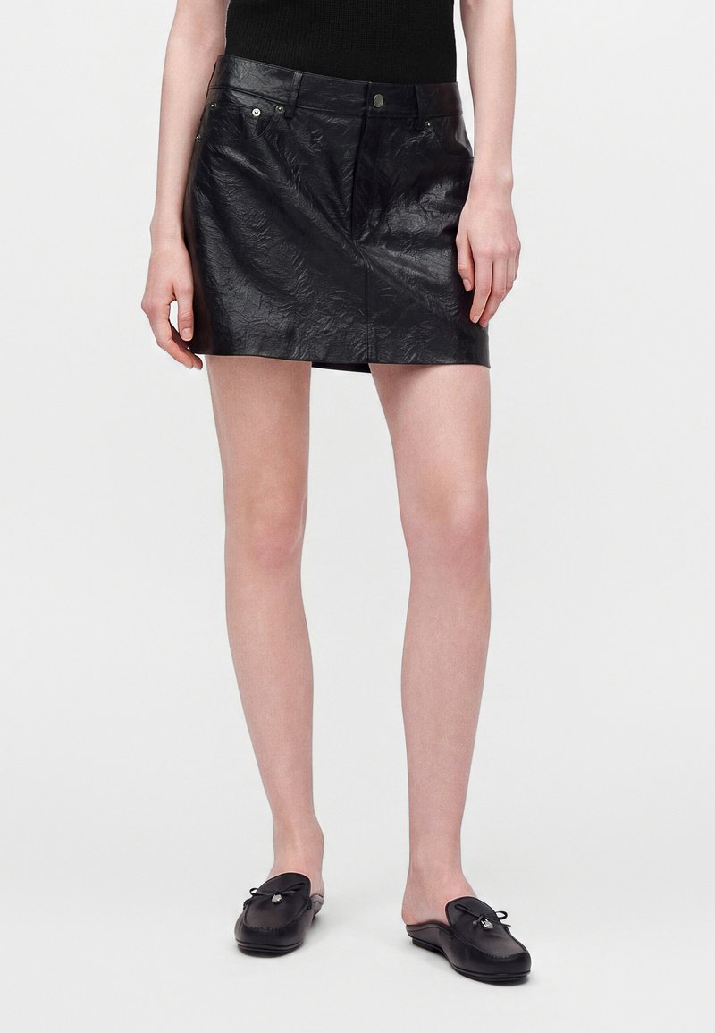 Personne portant une mini-jupe en cuir texturé noire, un haut noir sans manches et des mocassins noirs sans lacets, debout devant un fond uni.
