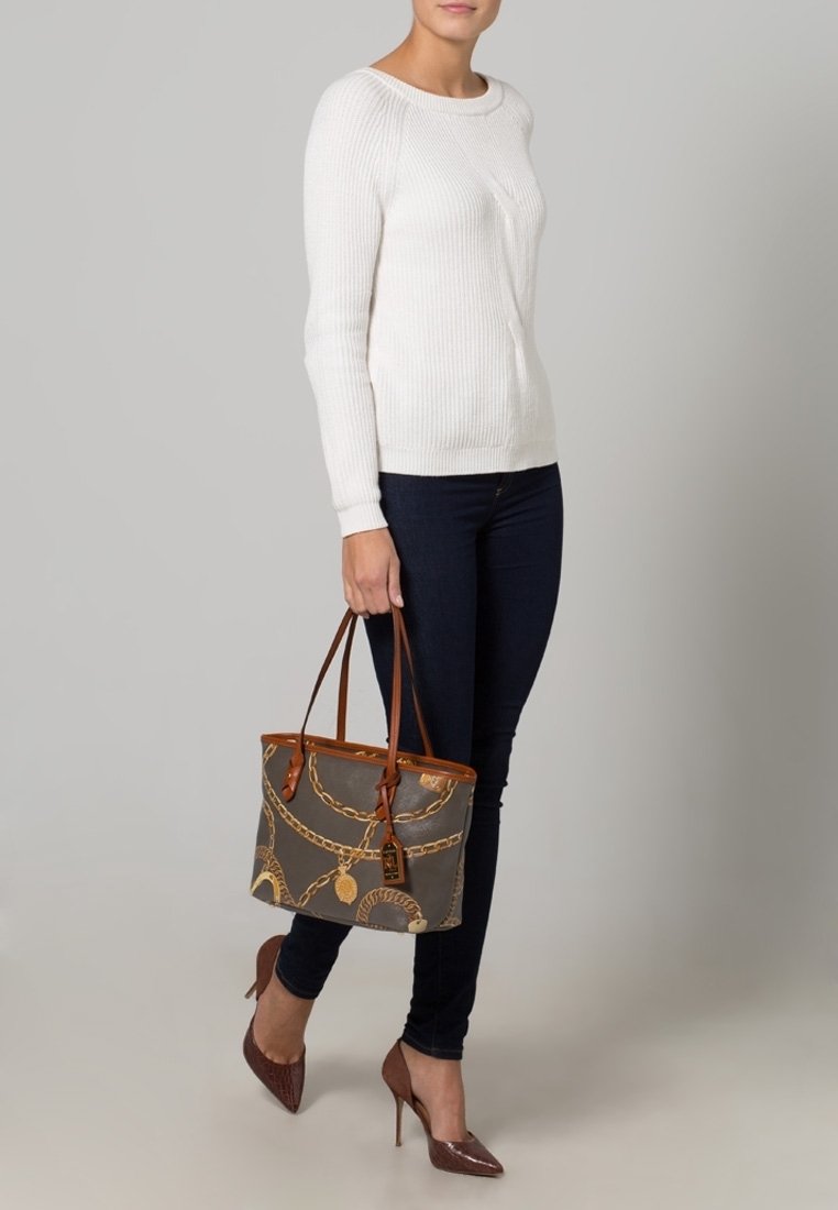 Femme portant un pull blanc, un jean skinny foncé, des talons marron, tenant un sac à main gris et marron à motifs avec des chaînes.