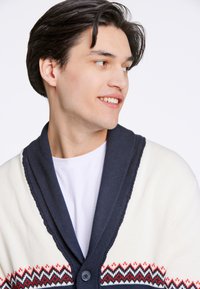 Stickad kofta i krämfärgad med en marinblå sjalkrage. Har ett geometriskt mönster i rött och blått vid fållen och en knappstängning.