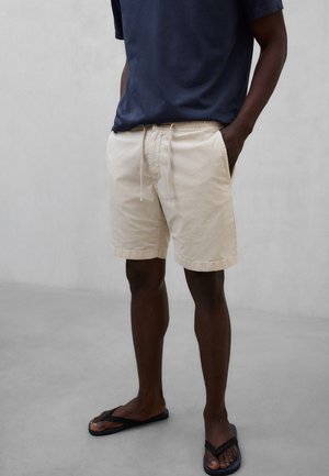 Hombre con pantalones cortos beige con cordón, camiseta azul marino y chanclas negras, de pie sobre un suelo gris liso.