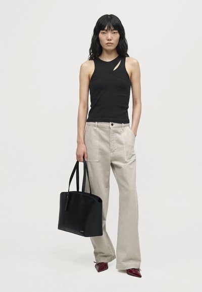 Une femme porte un haut noir sans manches, un pantalon beige ample, des chaussures bordeaux et un cabas noir, devant un fond uni.