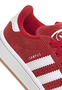 Sneaker in suede rosso con accenti in pelle bianca, suola in gomma e tre strisce bianche con il testo "CAMPUS" sul lato. Dettagli testurizzati visibili.