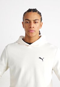 Hvid hættetrøje lavet af blødt stof med en kængurulomme og et sort Puma-logo på venstre bryst. Har snøring i hætten og en afslappet pasform.