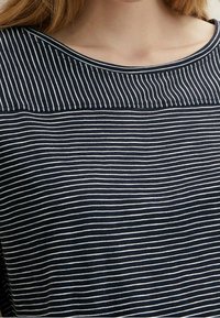 Kvinna iklädd en marinblå och vit randig topp med vid halsringning och horisontella ränder på bröstet och kroppsdelen.