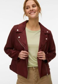 Femme souriante, portant une veste bordeaux zippée sur un t-shirt vert clair et un pantalon beige, tenant les revers de la veste avec les deux mains.