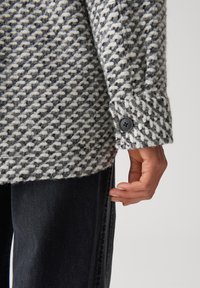 Grau und weiß strukturierter Wollmantel mit einem Hahnentrittmuster, ausgestattet mit einem geknöpften Ärmel und einer sichtbaren Hand, die auf dem Stoff ruht.