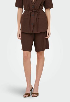 Kvinde iført en brun kortærmet blazer med bælte, matchende knælange shorts og brune åbne høje hæle sandal.