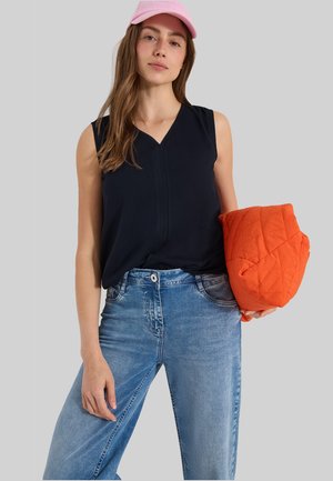 Femme portant un haut noir sans manches, une casquette rose, un jean bleu, tenant une veste matelassée orange pliée devant un fond gris.