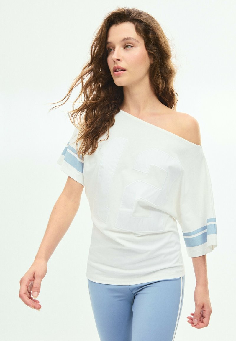 Femme aux longs cheveux ondulés portant un haut blanc épaules dénudées avec des rayures bleues et un pantalon bleu clair ajusté, regardant sur le côté.