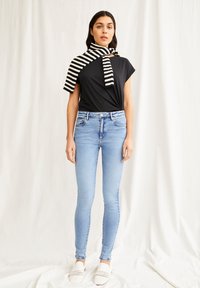 Camiseta negra, diseño anudado, bufanda a rayas blancas y negras doblada, jeans ajustados azul claro, zapatos slip-on blancos de tejido, de pie sobre un fondo liso.
