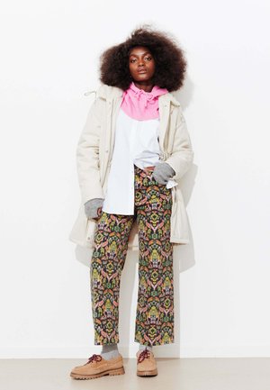 Parka beige, pull rose, chemise blanche, et pantalon large à motifs verts, roses et marron. Chaussures marron avec lacets. Gants gris.