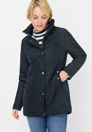 Woman wearing a navy blue buttoned jacket over a striped turtleneck sweater, with one hand in her pocket, looking down.