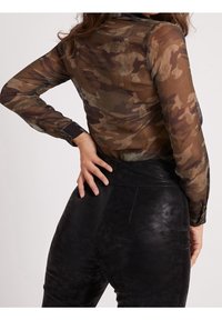 Langarmige, doorzichtige camouflage blouse in bruin en groen, gecombineerd met hoge, zwarte faux leren broek. Bevat een knoopmanchet.