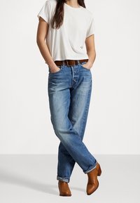 Lekka biała koszulka, niebieskie jeansy o prostym kroju, brązowy pasek i brązowe botki za kostkę. Jeansy są lekko wyblakłe z podwiniętymi mankietami.