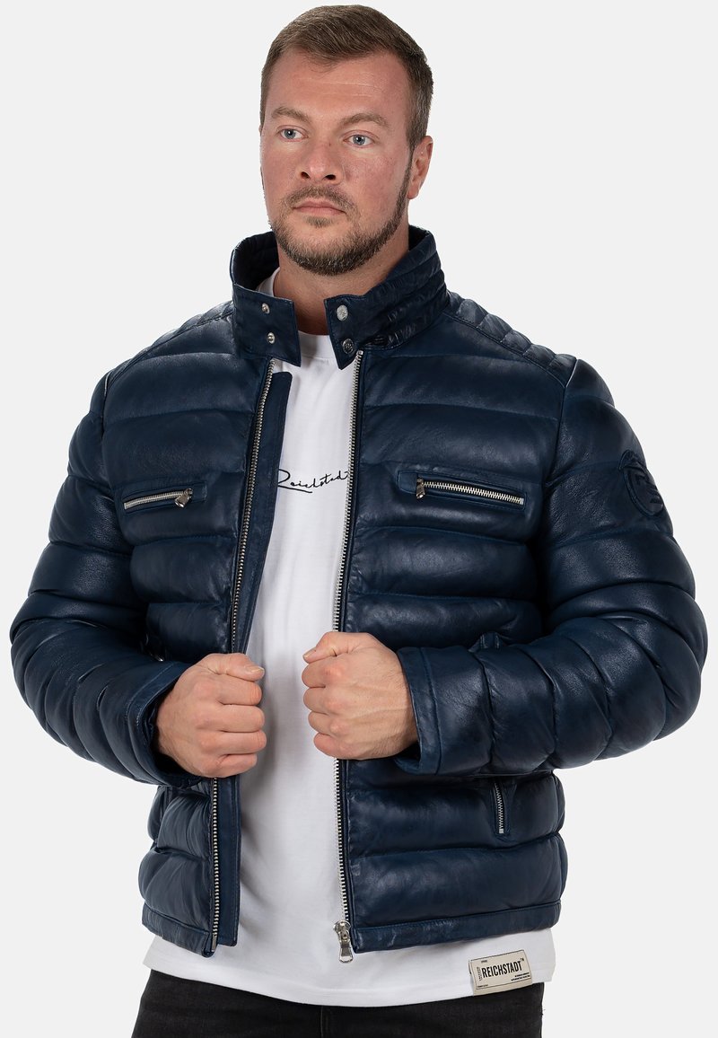 Giacca puffer navy in pelle con colletto alto, zip frontale e due tasche sul petto con zip. Presenta dettagli a costine sulle maniche.
