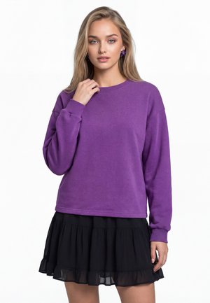Kvinde med langt blondt hår iført lilla sweater, sort lagdelt nederdel og lilla stjerneformede øreringe, stående mod hvid baggrund.
