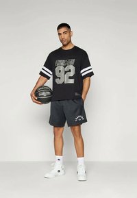 Μαύρο αθλητικό μπλουζάκι με την εκτύπωση "CHAMPION 92" σε γκρι, συνδυασμένο με γκρι αεροδιαπερατά σορτς και άσπρα αθλητικά παπούτσια. Κρατάει μια μαύρη μπάλα μπάσκετ.