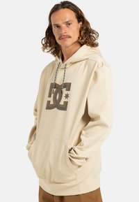 Beżowa bluza z kapturem z dużym logo DC w ciemnym brązie, regulowanym sznurkiem oraz przednią kieszenią kangurką. Gładka tekstura, luźny krój.