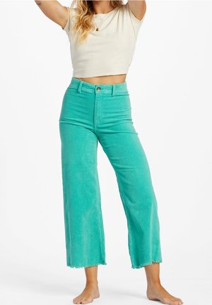 Kvinde iført turkise bukser med vide ben og en kort cremet T-shirt, stående barfodet med armene hævet mod en hvid baggrund.