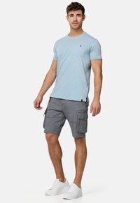 Helles blaues T-Shirt mit Rundhalsausschnitt und Tasche; graue Cargo-Shorts mit Seitentaschen; weiße Turnschuhe, stehend vor einem schlichten Hintergrund.