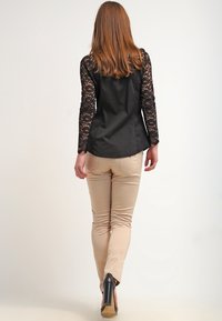 Svart blus med spetsärmar, figurnära design, kombinerad med beiga byxor och svarta klackar. Synlig ryggdetaljering och texturerade spetsaccenter.