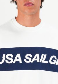 Man wearing white sweatshirt with a navy blue stripe and bold white text partially reading "USA SAIL GR".