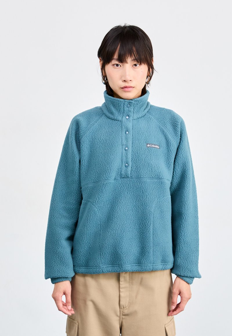 Pull-over en polaire bleu avec un col haut, une fermeture à boutons-pression et des manches raglan. Surface texturée et une poche sur la poitrine avec un détail de logo.
