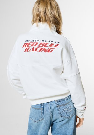 Persoon met blond haar die een witte Red Bull Racing trui draagt en blauwe spijkerbroek met Red Bull Racing borduurwerk op het achterzakje.