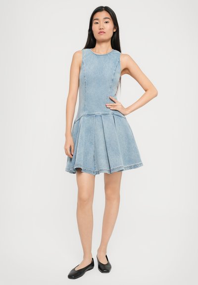 Asian woman standing with hand on hip, wearing sleeveless light blue denim dress with flared skirt and black flats, white background.